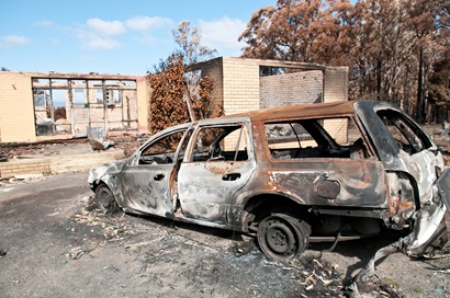 A burnt-out station wagon in front of a burnt-out dwelling, with only some walls still standing