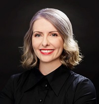 A woman with silver hair and red lipstick, smiling at the camera and wearing a black buttoned up top.