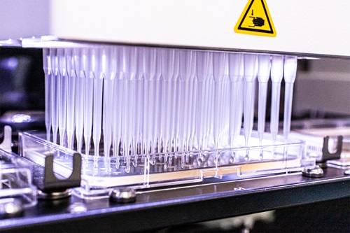 A close up of laboratory equipment with purple backlighting. It is a range of transparent pipettes accompanied by a yellow hazard icon.