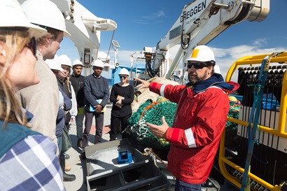 Man in a red coat instructs a group of students on the back deck of a ship.