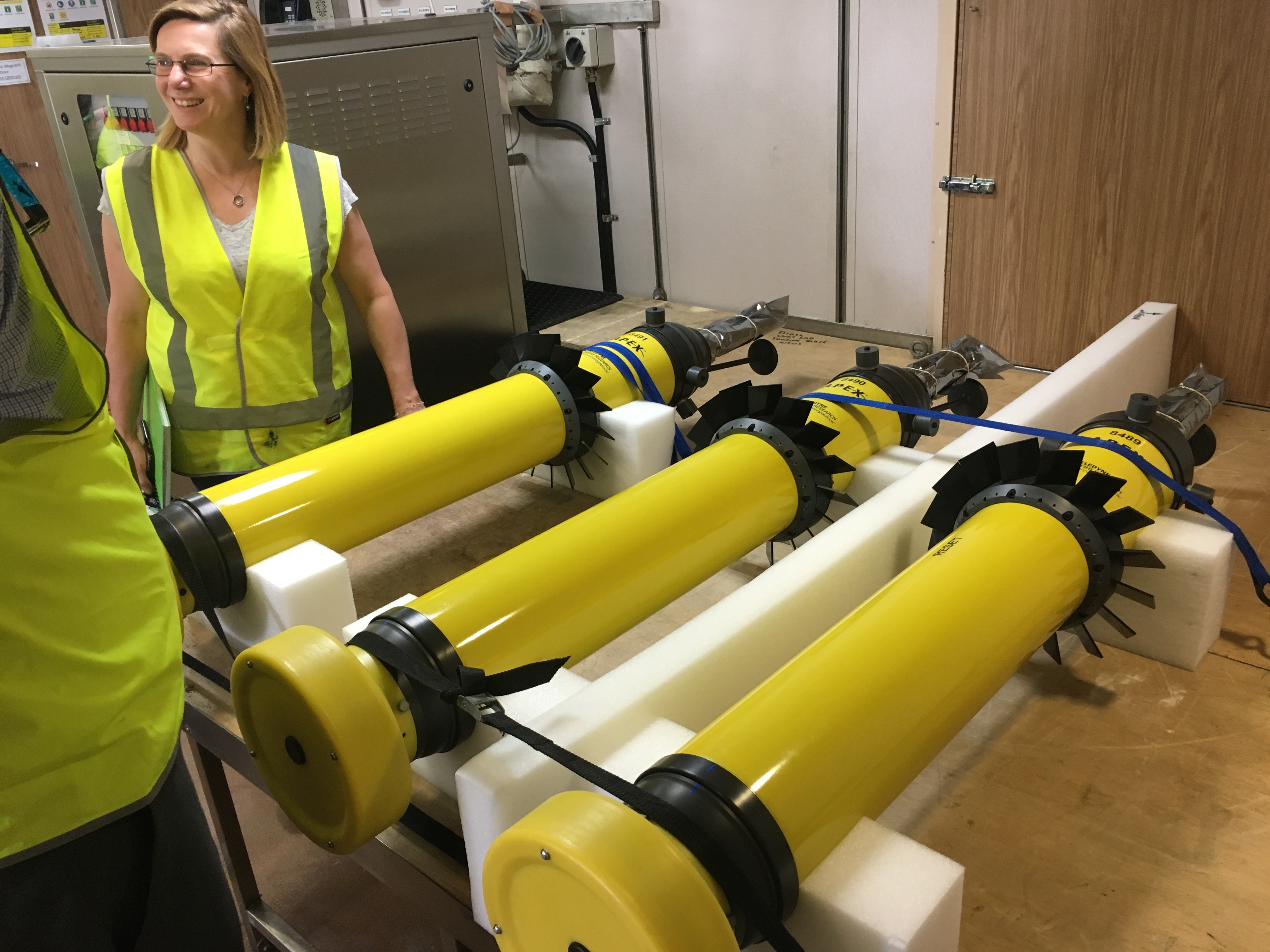 A row of bright yellow Argo floats sit on a bench with a woman in a yellow hi-vis vest in the background.