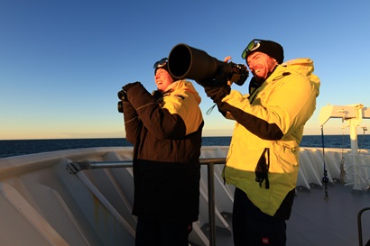A smiling man in a yellow raincoat holds a very large camera while a woman standing nest to him holds binoculars and smiles.