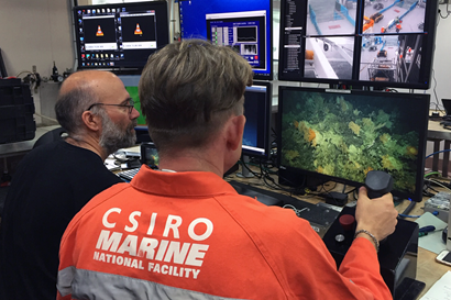 A man in an orange work suit with CSIRO Marine National Facility written on the back holds a joystick and stares at a screen showing an underwater image.