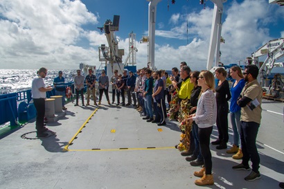 A group of people, some holding wreaths, stand in a semi-circle around a person in uniform on the back deck of a ship.