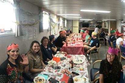 People in Christmas hats sit around tables in the mess of a ship.