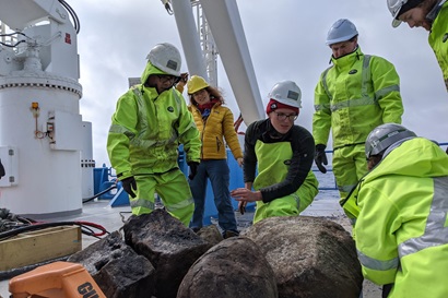Six people in hi-vis wet weather gear look at rocks on the deck of a ship.