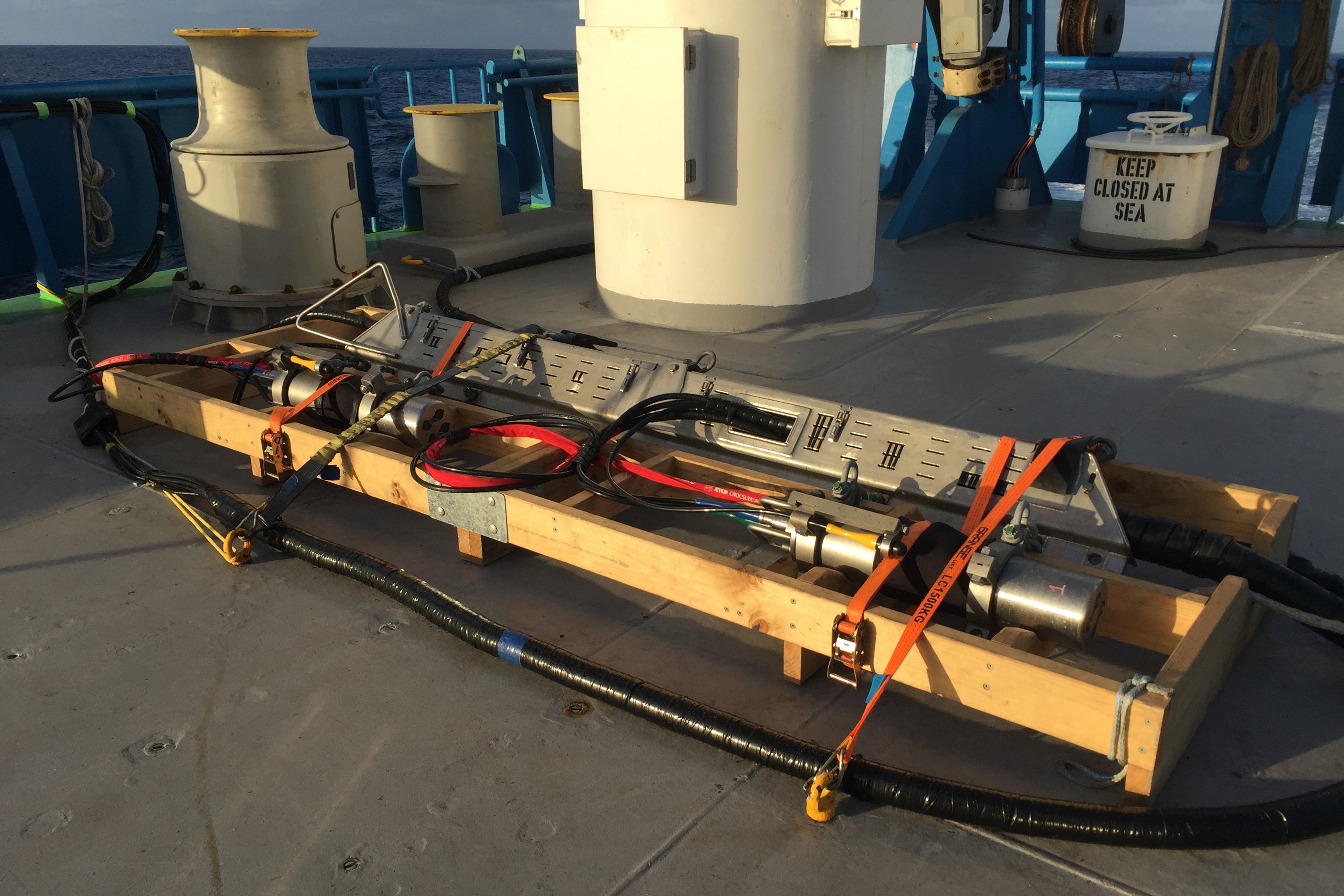 A large silver-coloured piece of scientific equipment sits in a rectangular wooden frame on the deck of a ship.