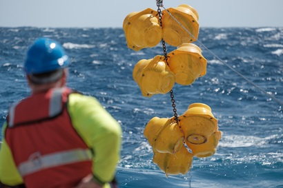 A person in a blue hard hat and red life preserver looks at a collection of yellow buoys being lifted from the ocean.