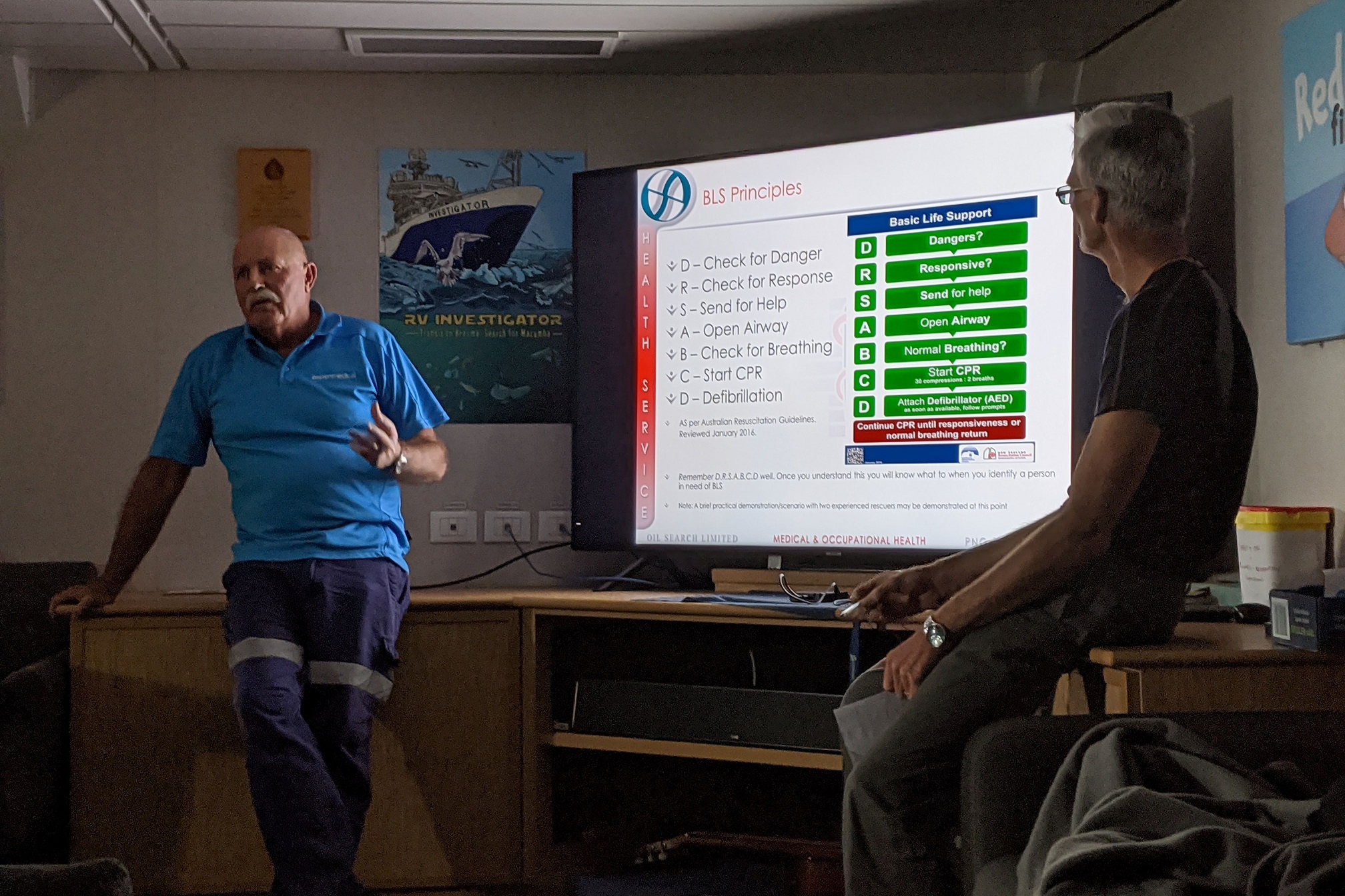 Two people sitting either side of a screen showing a first aid presentation.