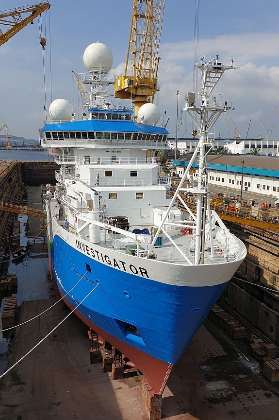 A large blue and white ship sits on blocks in a dock that has been emptied of water.