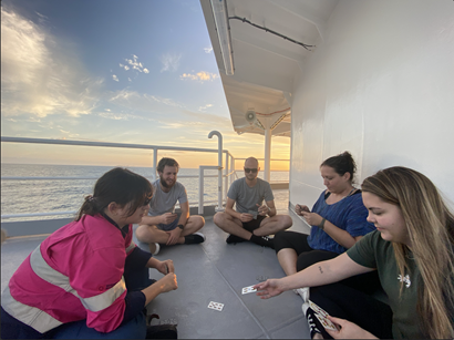 A group of people sitting on the deck of a ship at sea playing cards.