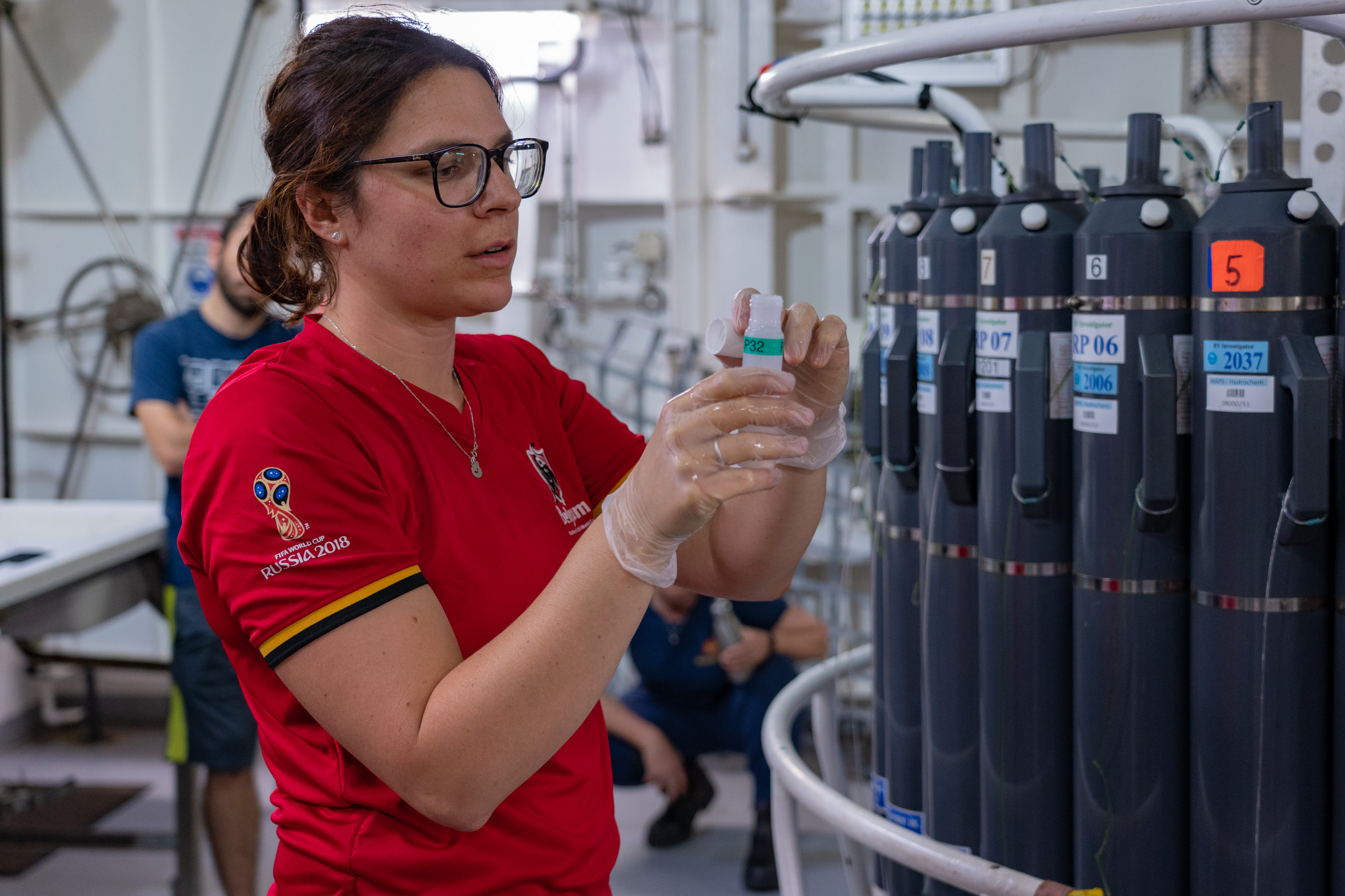 A person in a red t-shirt wearing clear gloves handles a small vial next to a large piece of scientific equipment with rows of tall grey bottles around it.