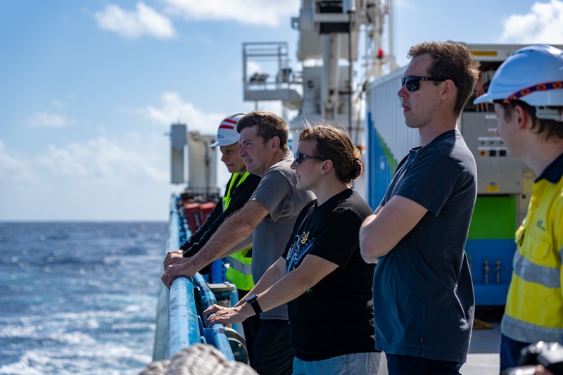 A group of people along the railing of a ship stare out to sea.