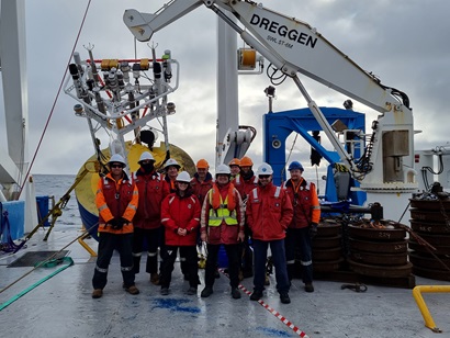 A group of people in wet weather gear and hard hats stand in front of a large piece of scientific equipment on the back deck of a ship.