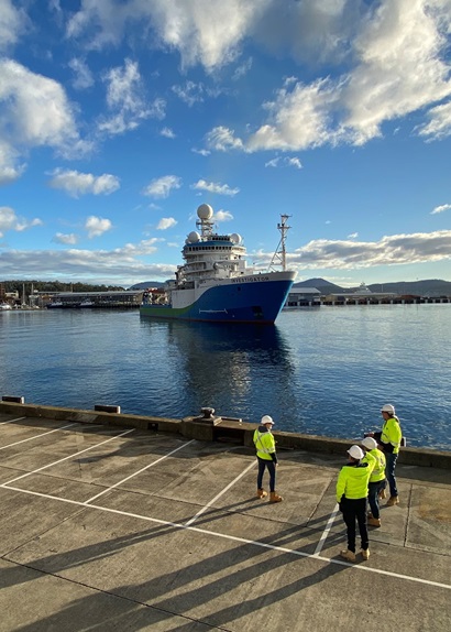 A blue and white ship is watched by four people in high vis clothing and hard hats as it pulls into port.