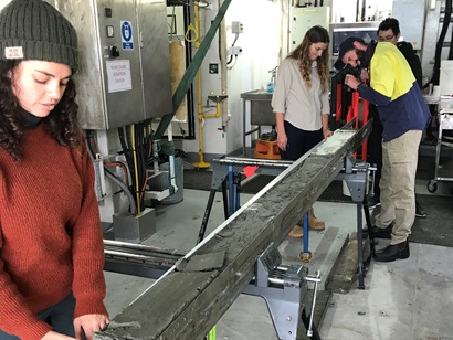 A group of people look at a long metal tray filled with grey mud.