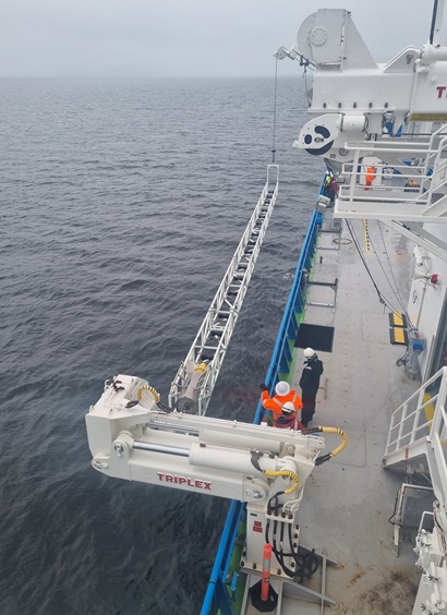 Two cranes on a ship suspend a long frame that looks like a girder over the ocean.