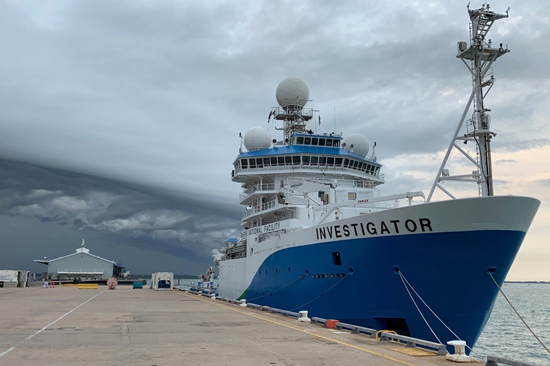 A blue and white ship tied up next to a wharf with a stormy sky behind.