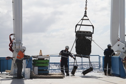 Three men in overalls and hard hats dragging a large metal bag of rocks onto the back deck of a ship.