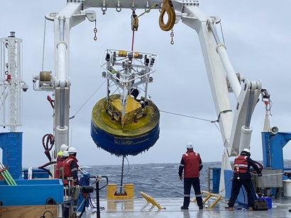 A large yellow and blue piece of scientific equipment shaped like a cork hangs from the A-frame on the back of a ship at sea.
