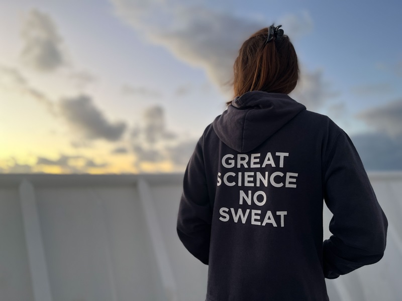 A person in a blue hoody with words 'Great science, no sweat' on the back stands on the deck of a ship.