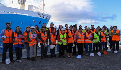 A large group of people in orange and yellow hi-vis vests and some with hard hats stand in front of a ship.
