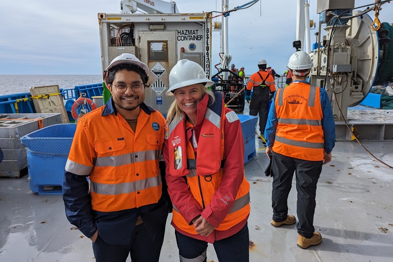 Two people in hi vis and hard hats stand on the deck of a ship.