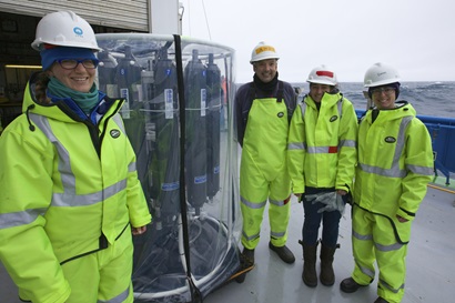 Four scientists in cold weather gear stand on the deck of a ship next to a large circular scientific instrument.