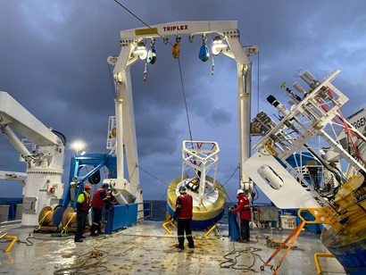 The back deck of a ship at sea showing a large piece of scientific equipment hanging from a crane.