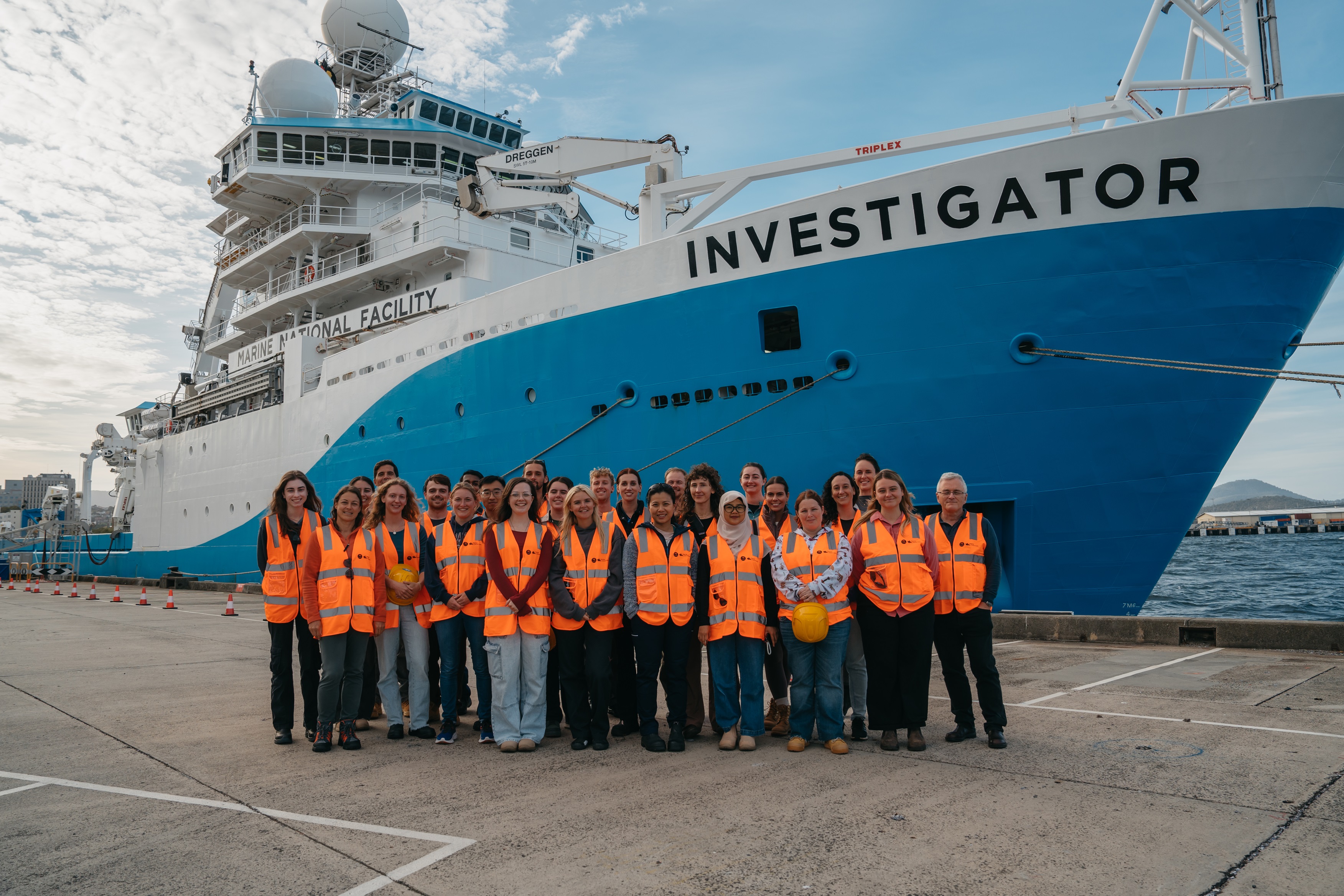 A groupf or people wearing hi-vis vests standing on a wharf in front of a large blue and white ship with Investigator written on its bow.