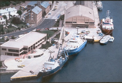 Three vessels alongside a wharf with various buildings next to the wharf.