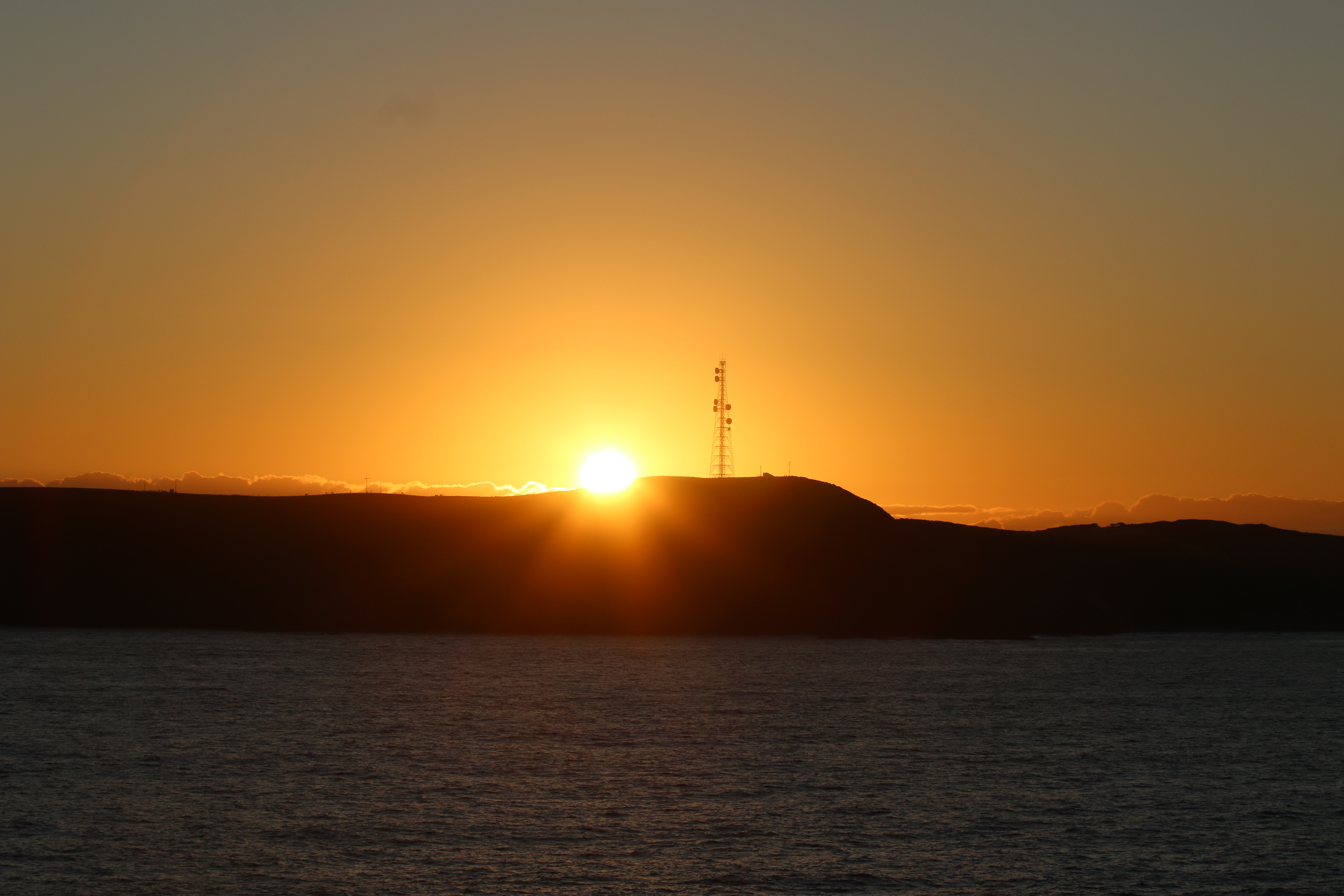 The sun rising over a headland with a tall twoer on it viewed from the ocean.