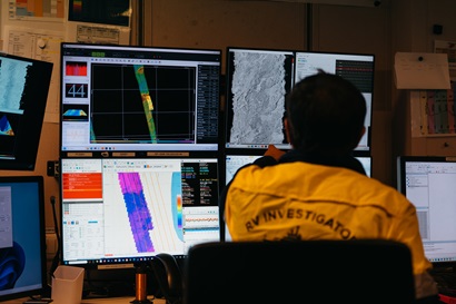 A person in a high vis shirt sitting in front of a bank of computer monitors.