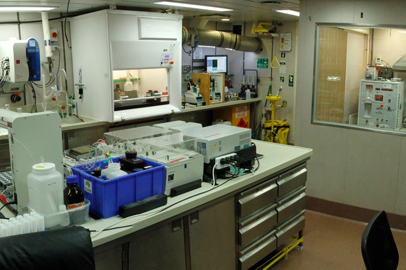 A scientific laboratory with a fumehood in the background and bench covered with scientific instruments in the foreground.