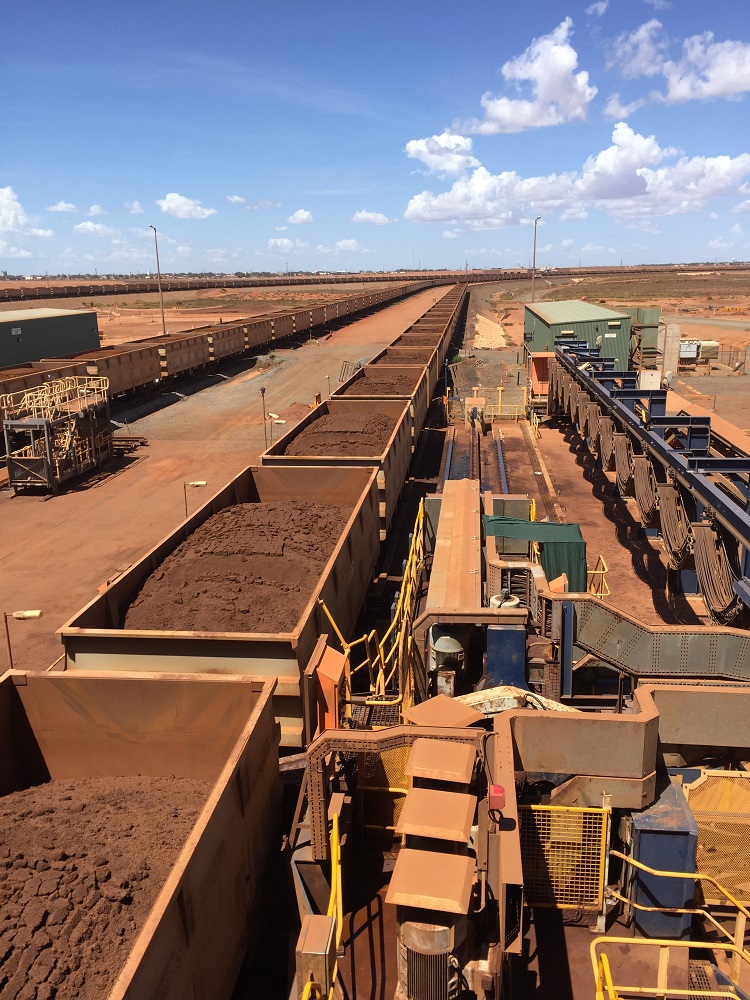 Long chain of open rail carriages filled with rust-coloured iron-ore rock winding into outback landscape