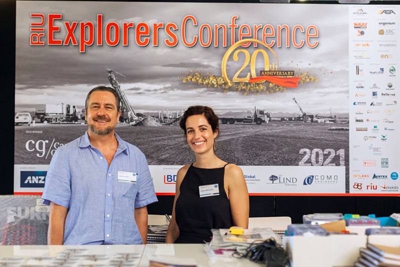Man and women standing in front of a desk and conference backdrop celebrating  RUI Exploreres conference 20th anniversary