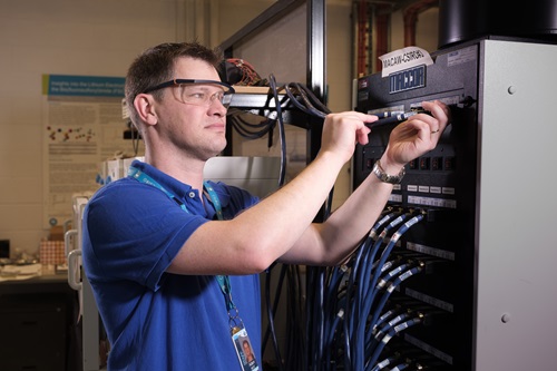 A man in blue t-shirt and safety glasses works with a screw driver on a tall grey box-like piece of equipment with a series of blue cables plugged into it.