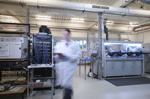 A blury image of a scientist in a lab coat walking through a lab with large battery testing equipment in background