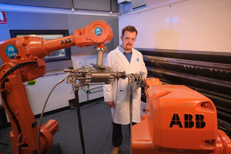 A man in protective glasses and lab coat stands next to machinery. 