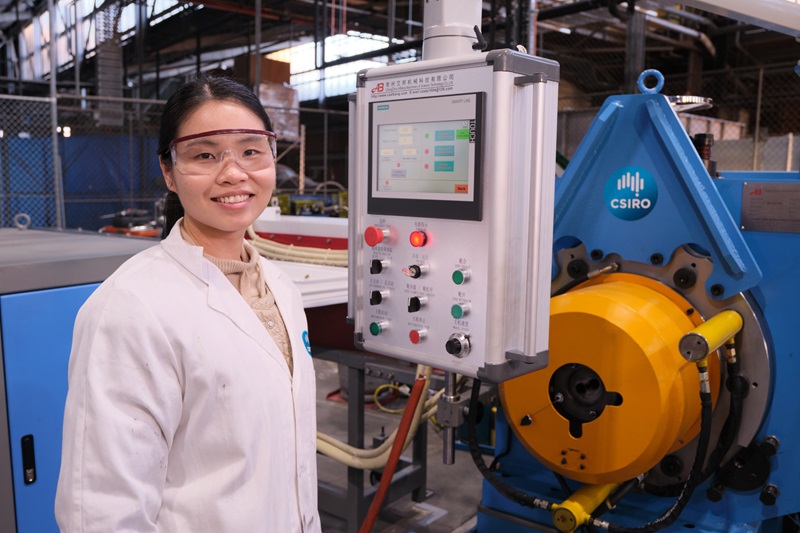 A woman in wearing protective gear stands in front of a machine at a factory. 