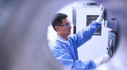 Scientist in a lab gown, gloves and safety glasses turning a dial on a 150L stirring tank bioreactor which is used for mammalian cell culture to produce recombinant protein for research.