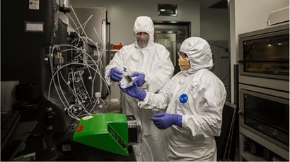 two researchers in white hazmat suits and blue gloves in a lab handling samples