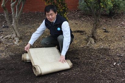 Image of Dr Malcolm Miao crouching down holding a the linseed matting. He's outside crouching down surrounded by trees. The matting is rolled up and is approximately 1 foot in width