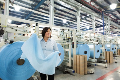 Lady stands in large fabric manufacturing plant in front of rolls of light blue felt fabric. She is holding a large piece of the fabric. 