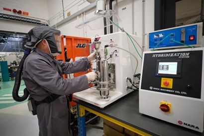 A researcher in a grey full PPE body coverall, including respiratory mask and gloves, works with industrial metal powder processing equipment in a lab environment. 