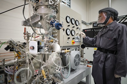 a researcher in a grey PPE coverall, including respiratory mask and gloves, works with a large industrial metal powder processing machine in a lab environment. You can see lots of plastic tubes and fans and a metal funnel attached to a large beige machine covered in various dials and switches. 