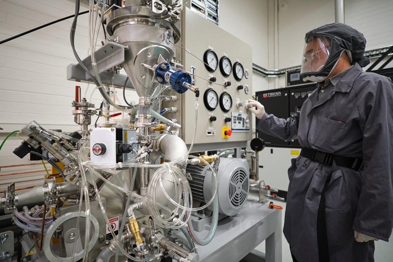 a researcher in a grey PPE coverall, including respiratory mask and gloves, works with a large industrial metal powder processing machine in a lab environment. You can see lots of plastic tubes and fans and a metal funnel attached to a large beige machine covered in various dials and switches. 