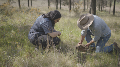 A scientist and a man are kneeling on the ground digging