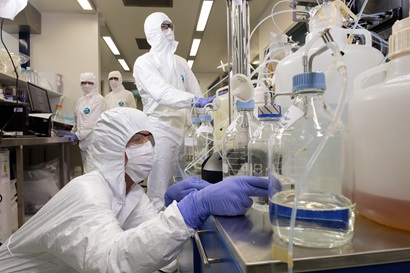 Researchers in white hazmat suits working in a lab. one researcher in foreground is bending down looking in a vessel