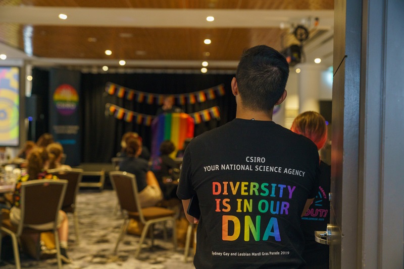 Young man stands facing away, listening to a speaker on a stage surrounded by rainbow flags and the rainbow CSIRO logo. His tshirt back print says CSIRO, your national science agency: Diversity is in our DNA.